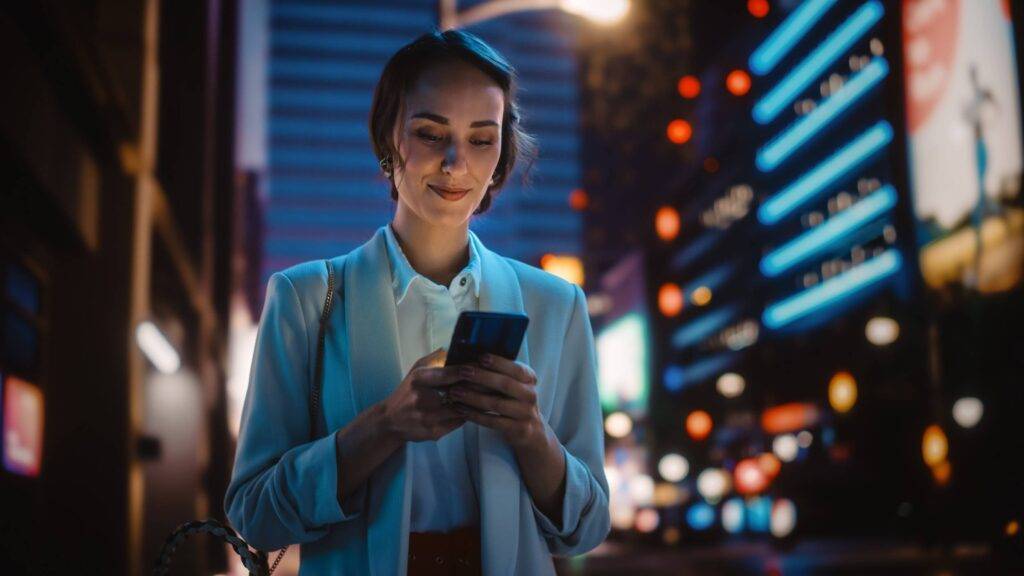 Beautiful Young Woman Using Smartphone Walking Through Night City Street Full of Neon Light. Smiling Thoughtfully Female Using Mobile Phone, Posting Social Media, Online Shopping, Texting.
